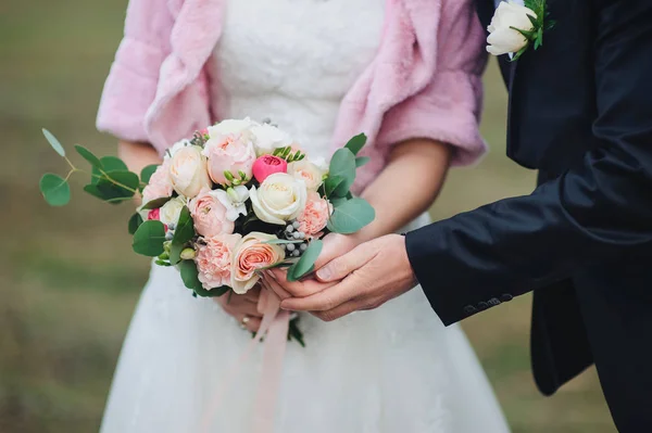 Young bride in pink fur coat holds a wedding bouquet in hands, the groom hugs her from behind. Autumn weather.