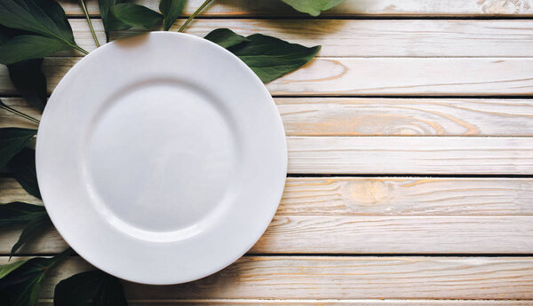 Empty white plate on light gray rustic wooden background with green leaves of peony. Top view, copy space, close up.