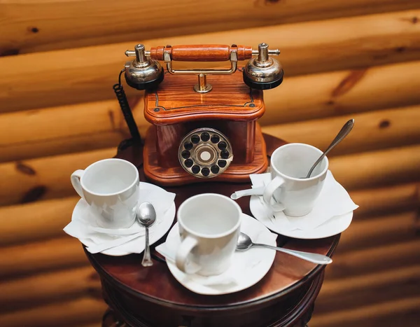 An old, antique, rarity telephone with a disk dialing set is standing on the table near empty white cups on a wooden background. Close up.