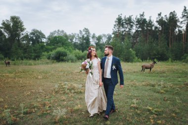 Gelin ve damat birbirine yaslanmış yaz orman glade yürüyorduk. Gelin buketi ve taze çiçek çelenk ile.