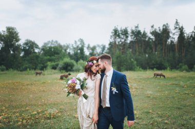 Gelin ve damat birbirine yaslanmış yaz orman glade yürüyorduk. Gelin buketi ve taze çiçek çelenk ile.