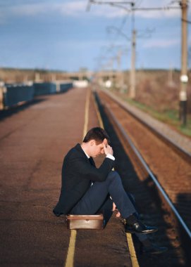 Genç bir ofis işçisi bir bavul üzerinde oturur ve üzücü. Bir platform, gün batımı, demiryolu parkuru.
