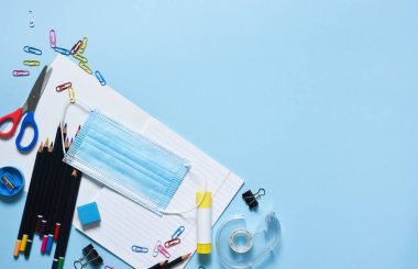 Mavi arka planda okul malzemeleri ve maske. Okul aksesuarlarının yerleşimi. Okula dönelim. Mesafe öğreniyor. Tahtada tıbbi maske var. Karantina eğitimi.