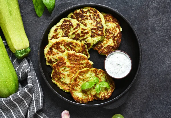 Zucchini pancakes with sauce and garlic in a frying pan on a black background. View from above.