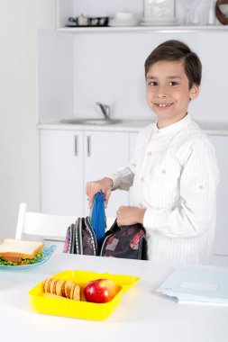 Bir okul çocuğu okula bir sırt çantası toplar. O bir kalem ve bir kutu öğle yemeği bir okul çantası koyar,