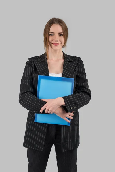 年轻成功的女人在一个严格的深色西装与一个蓝色的文件在手看着相机和微笑 — 图库照片
