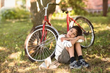 Oğlan bir elma bahçesi veya ormanında yere oturur. Bir ağaç yakınındaki bir şey hakkında düşünmeye o hayalleri standları bir kırmızı bisiklet ve kitap