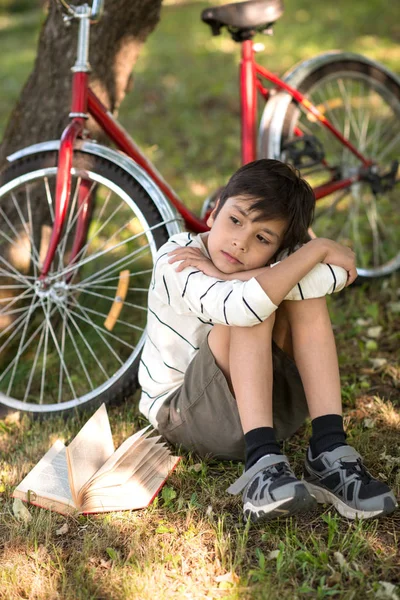 Oğlan bir elma bahçesi veya ormanında yere oturur. Bir ağaç yakınındaki bir şey hakkında düşünmeye o hayalleri standları bir kırmızı bisiklet ve kitap