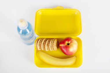  Öğle yemeği ile sarı plastik kutu ve okul için su şişesiyle yalan beyaz bir masaya. Bir muz, elma ve bisküvi kutusuna vardır, 