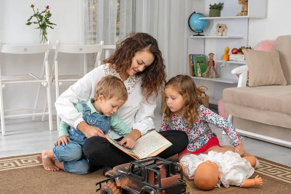 Genç Anne veya dadı ile küçük çocuk, bir erkek ve bir kız evde odada halı katta oturmak ve bir kitap okumak bir oyuncak araba ve bir bebek ile oyna
