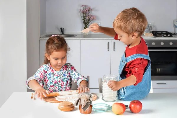 Little girl kneads dough on kitchen table at home with delight. Her younger brother is sitting on the table and pouring flour over the dough