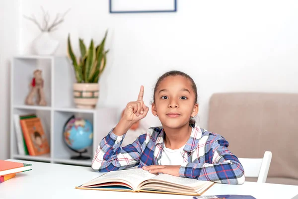 The little mulatta girl in a plaid blue shirt is sitting at the table in the room and doing homework.  She shows an index finger up asking for this gesture of attention