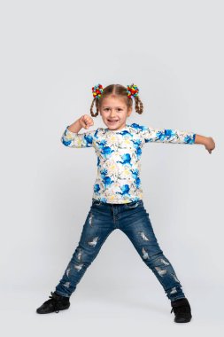 Tam uzunlukta stüdyo çekim küçük sarışın gülümseyen kız giyiyor renk gömlek ve gri arka plan üzerinde kot pantolon. O modern dans hareketlerini gösterir.