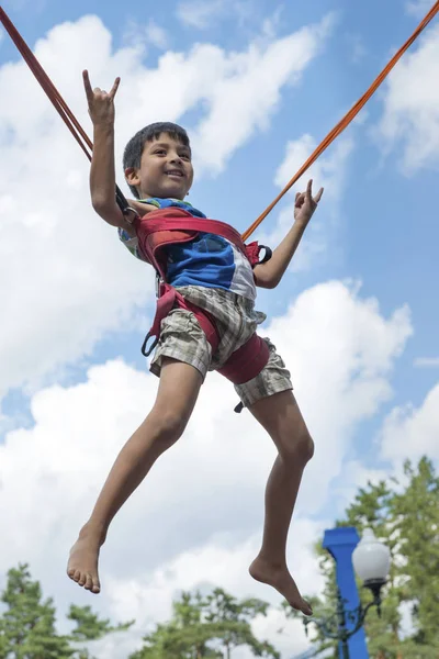 A little boy is jumping on a trampoline in the park.  He soars in the air against the blue sky and shows a gesture that he is doing fine