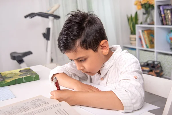 De school jongen zit aan tafel thuis en doet zijn huiswerk. — Stockfoto