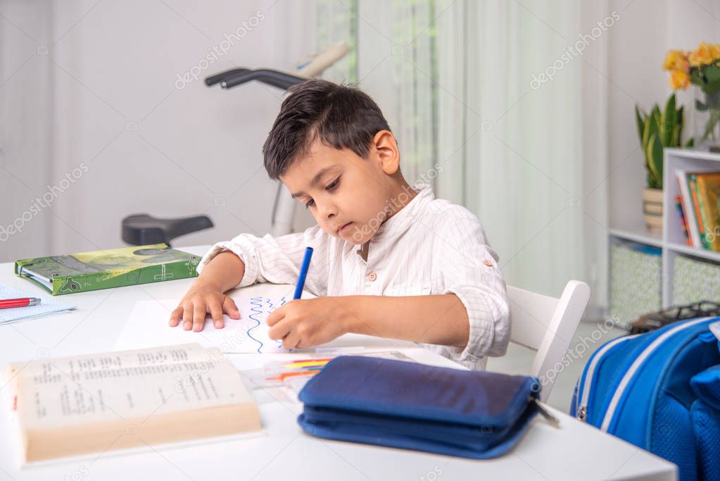 Waist up portrait of serious schoolboy  siting at the table at h