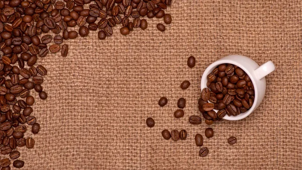 Coffee grains are scattered on a jute napkin.  Nearby is an overturned cup of coffee beans.Texture background. Copy space. The concept of cheerfulness and good mood.