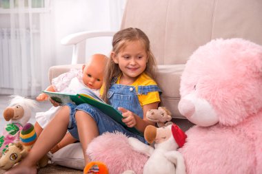 Yanında oyuncaklarıyla kitap okuyan küçük bir kızın fotoğrafı. 