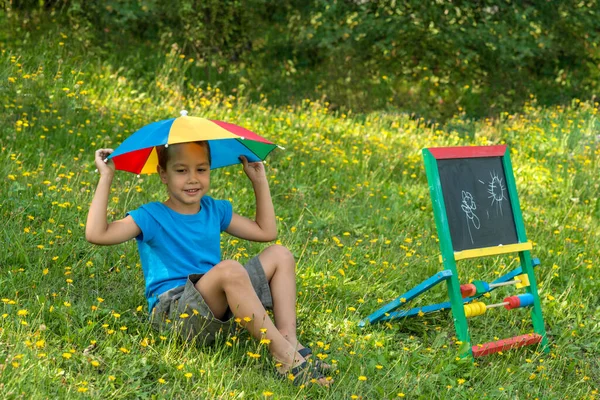 Shot of  a little  boy with sun umbrella sitting on the grass with yellow flowers in the garden on a sunny summer vacation day.   Concept of a happy childhood.