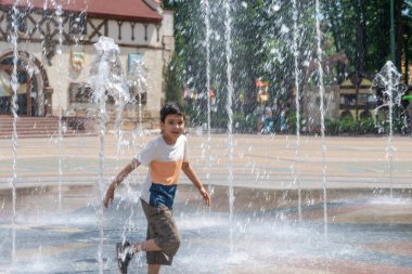 Sıcak bir yaz gününde şehirde bir parkta fıskiyede suyla oynayan genç bir çocuk.