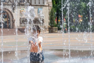 Sıcak bir yaz gününde şehirde bir parkta fıskiyede suyla oynayan genç bir çocuk.