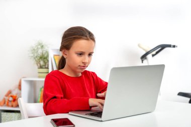 Kırmızı tişörtlü bir kız öğrenci odada dizüstü bilgisayarla bir masada oturuyor. O bir dizüstü bilgisayar kullanarak ödev yapıyor..