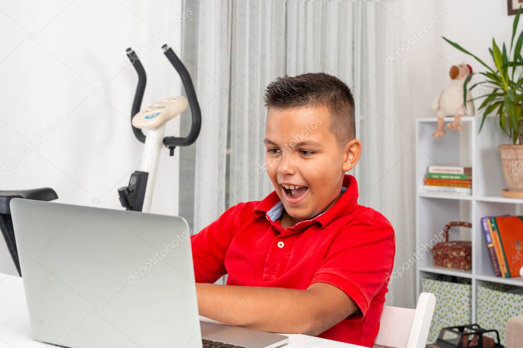 Shot of  schoolboy wearing red shirt  sitting at a table with a laptop in the room.  He  emotionally plays computer games or communicates on the Internet