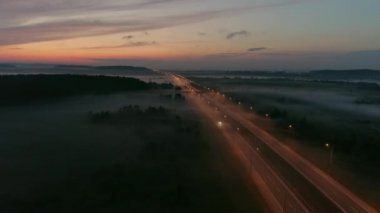 Gece otobanındaki araba sürüşünün hava görüntüsü. Işıklar parlıyor. Ormanın ve tarlanın kenarlarında. Sis tarlaların üzerinde. Şehir ufukta görünüyor. Güzel gün batımı ve gökyüzünde bulutlar