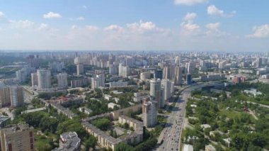 Araba trafiği olan modern yüksek binaların olduğu büyük şehir manzarası. Şehirde bir sürü yeşillik ve ağaç var. Yaz güneşli bir gün. Mavi gökyüzünde bulutlar var