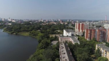 Nehrin yanında eski Sovyet yüksek binaları olan yerleşim yeri manzarası. Güneşli bir yaz günü. Evlerin arkasında bir gölet ve fabrika var. Arabalar caddelerden geçer. Kamera ağaçlara doğru iniyor.