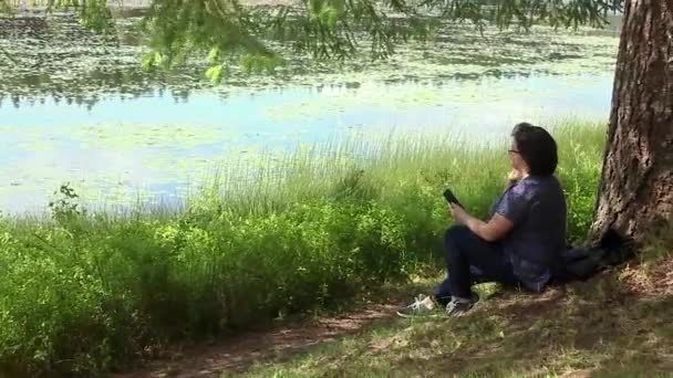 Ung kvinna med telefon under träd nära lake — Stockvideo