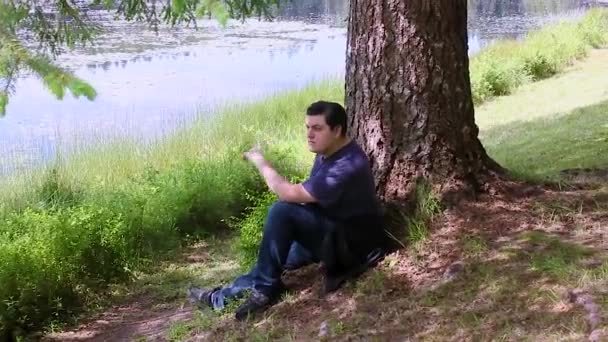 Un tipo sentado debajo de un árbol cerca de un lago — Vídeo de stock