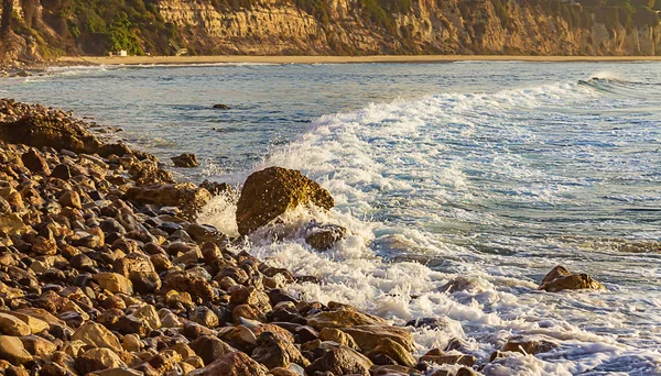 Foaming wave breaking on rocky shore across expanse of bay with cliff and sandy beach, — Stock Photo, Image