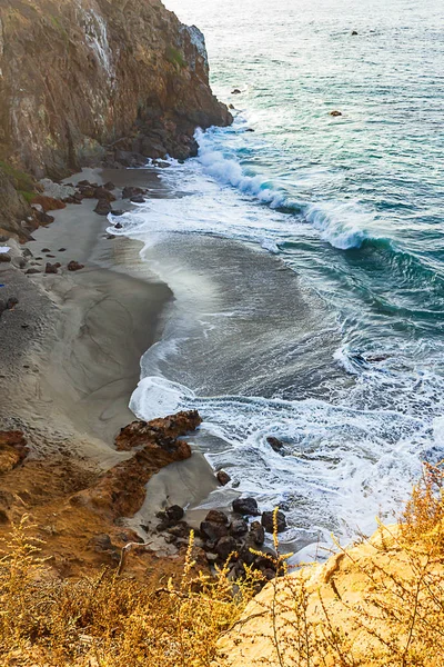 Rompiendo olas y senderos de espuma en una playa de arena oscura con rocas y borde del acantilado —  Fotos de Stock