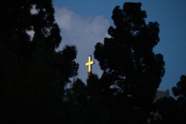 bulutlar ile mavi bir gökyüzünde parlayan beyaz bir haç çevreleyen siluetli ağaçlar