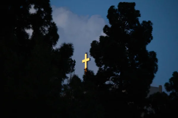 Árboles siluetas que rodean una brillante cruz blanca en un cielo azul con nubes — Foto de Stock