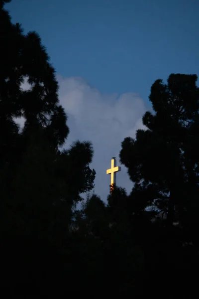 Árboles siluetas que rodean una brillante cruz blanca en un cielo azul con nubes — Foto de Stock