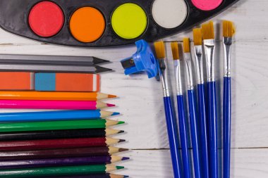 Beyaz tahta zemin üzerinde çizim araçları kümesi. Suluboya resimler, boya fırçaları, renkli kalemler, silgi ve masanda bileyici. Üst görünüm