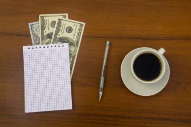 Bir fincan kahve, dolar, not defteri ve kalem ahşap masa üzerinde. Finansal planlama kavramı