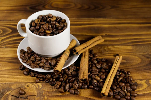 Granos de café en taza blanca y palitos de canela en mesa de madera —  Fotos de Stock