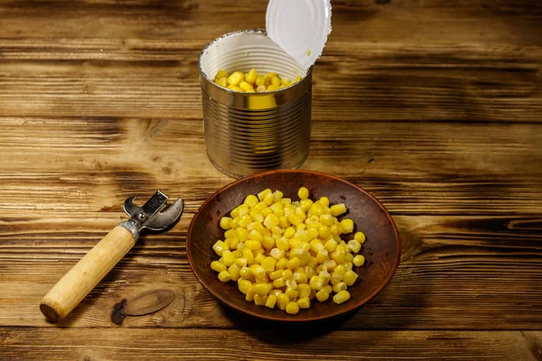 Open tin can and plate with corn and can opener on wooden table — Stock Photo, Image