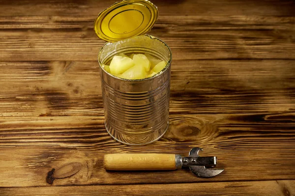 Opened tin can of pineapple pieces and can opener on wooden table