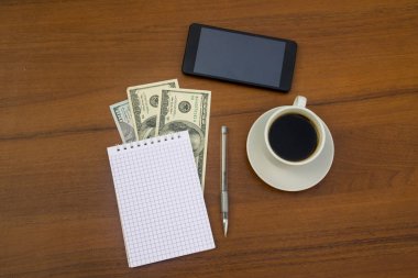 Bir fincan kahve, akıllı telefon, dolar, not defteri ve tahta masadaki kalem. Mali planlama kavramı