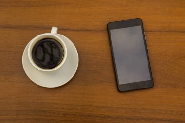 Ahşap masada bir fincan kahve ve akıllı telefon. Üst görünüm