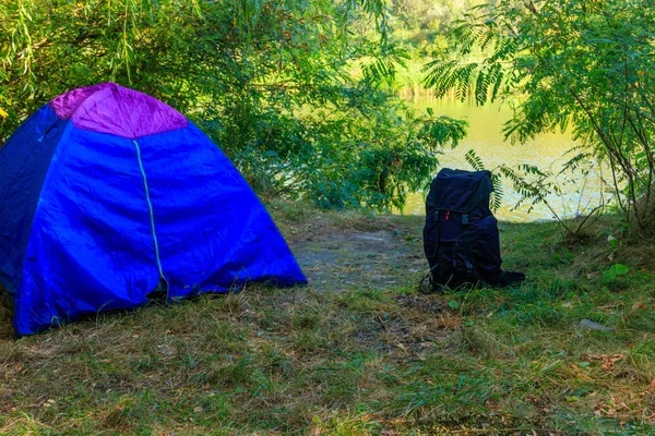 Mavi kamp çadırı ve sırt çantası bir nehir yakınında ormanda — Stok fotoğraf
