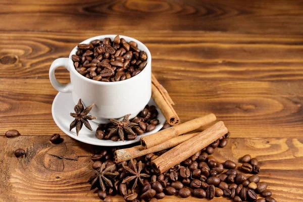 Granos de café en taza blanca, palitos de canela y anís estrellado en mesa de madera —  Fotos de Stock