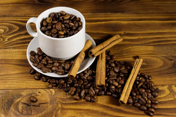 Granos de café en taza blanca y palitos de canela en mesa de madera —  Fotos de Stock