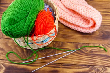 Hasır sepet, renkli örgü iplikleri, örgü iğneleri ve ahşap masa üzerine örülmüş eşarp.