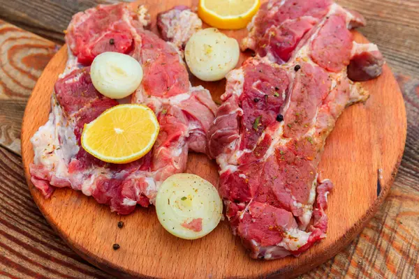 Carne fresca de cerdo cruda con especias, limón y cebolla sobre tabla de cortar de madera sobre mesa rústica —  Fotos de Stock