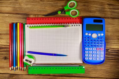 Okul kırtasiye malzemeleri seti. Boş not defteri, hesap makinesi, cetveller, kalemler, kalemler, makas ve tahta masada kalemdelen. Üst görünüm